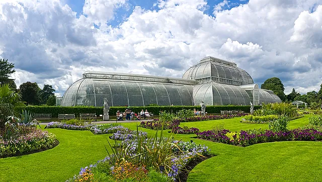 The curved glass and white iron Palm House beyond Victorian flower beds and lawn, under a sky of broken cloud