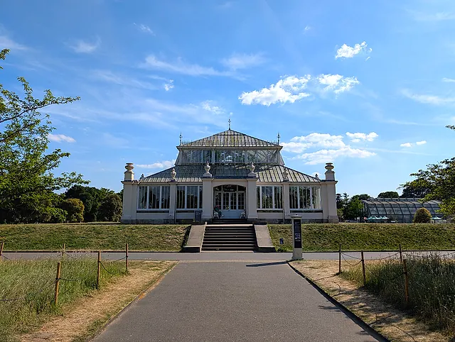 The Temperate House seen straight on from its approach path, with steps up to the central entrance and a blue sky